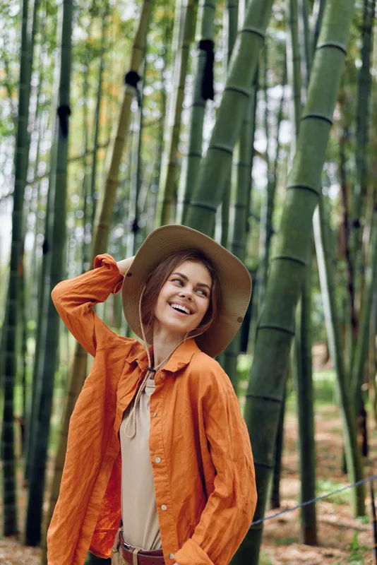 Woman smiling in hat among tall bamboo forest, nature portrait of a traveler wearing orange jacket and straw hat, candid outdoor moment showing eco tourism, adventure and joyful expression.