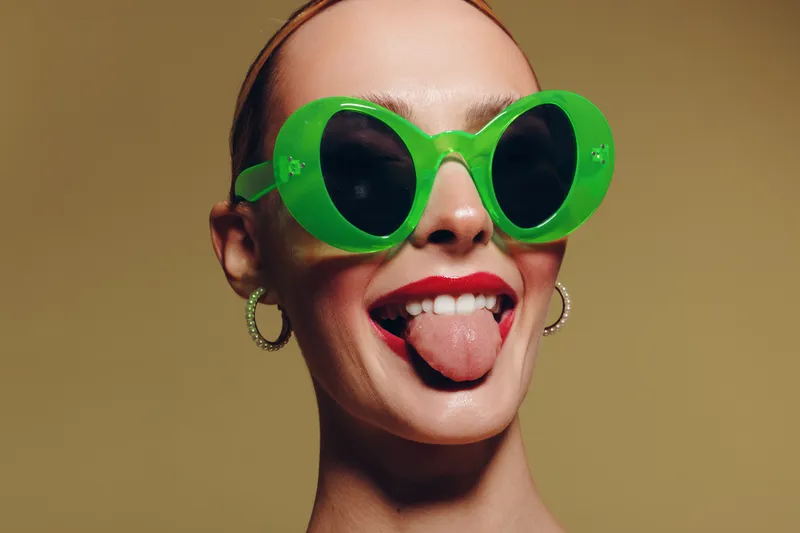 Young woman with bright red lipstick wearing large green sunglasses sticks out her tongue. Close-up portrait on beige background with playful and confident expression.