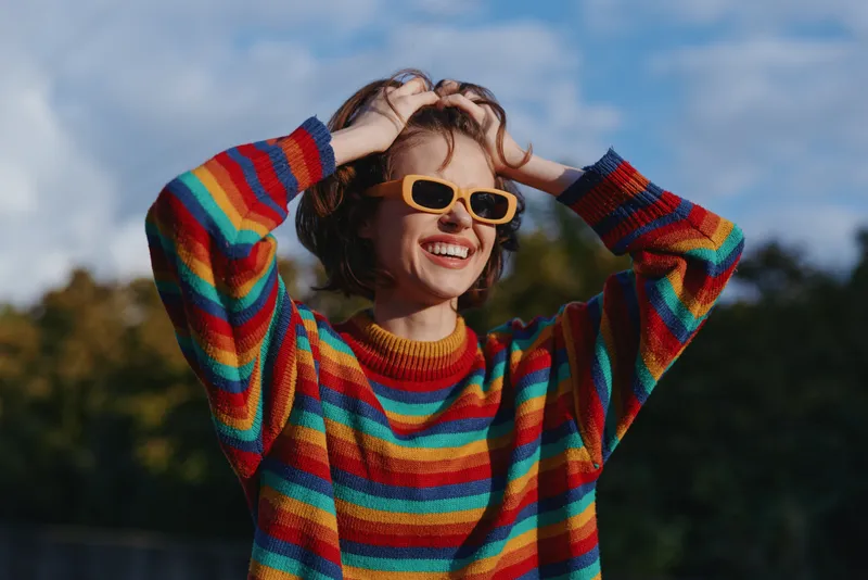 Woman in her 20s wearing a colorful striped sweater and yellow sunglasses, smiling with hands in hair outdoors in a park, casual outfit, joyful fashion lifestyle and warm autumn light.