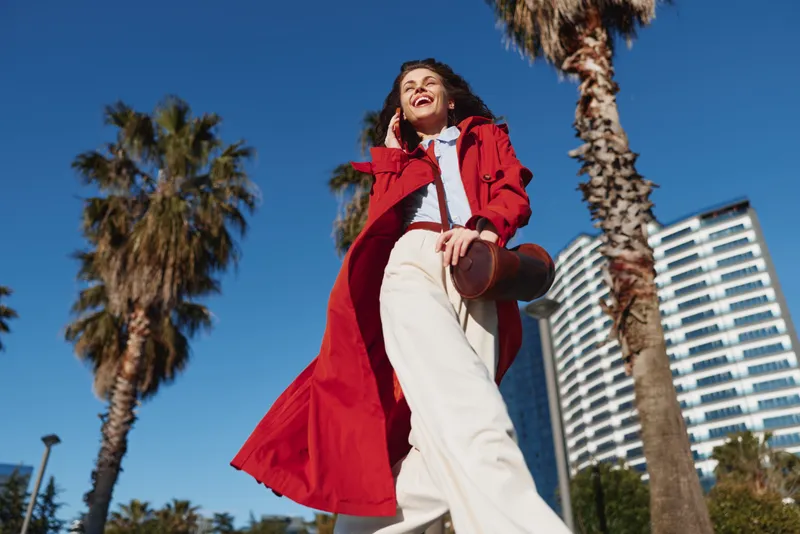 Young woman in a flowing red coat walks confidently outdoors on a sunny day with palm trees and modern buildings in the background, smiling joyfully.