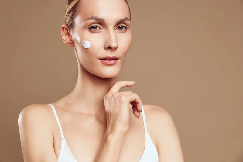 A young woman with clear skin applies white cream on her cheek, looking at the camera. The neutral background emphasizes skincare and beauty concepts.