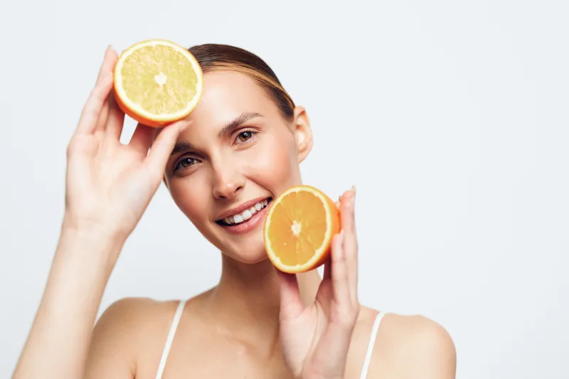 A smiling young woman holds two halves of a fresh orange near her face against a plain background. The concept highlights healthy lifestyle, natural beauty, and fresh fruit.
