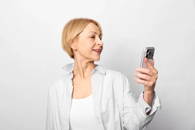 Smiling senior woman holding smartphone for a selfie, relaxed portrait in a white studio, soft light and calm mood, close framing highlights face expression and hands