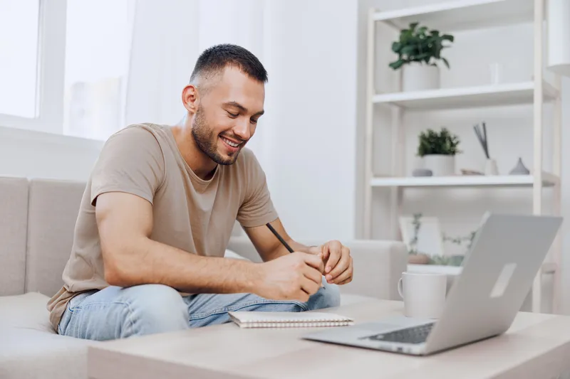 Cheerful man working from home, engaged in creative tasks with a laptop, casual attire, bright indoor setting, inspiring workspace atmosphere.