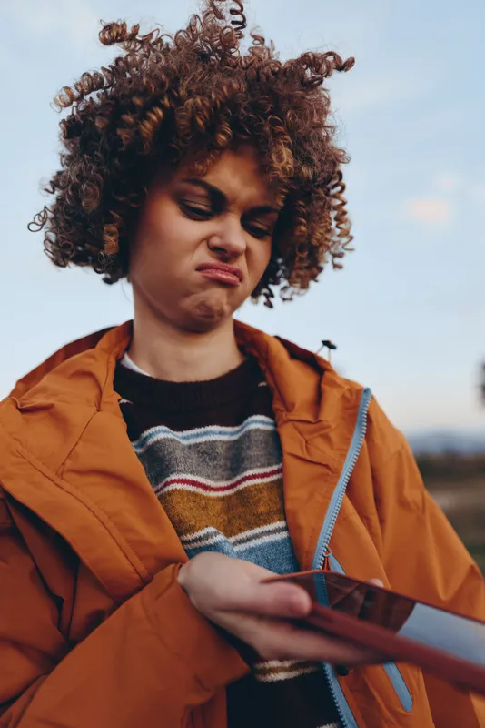 Woman with curly hair outdoors in an orange jacket holds a phone, autumn lifestyle portrait, thoughtful expression against a bright sky, outdoor moment in warm light.