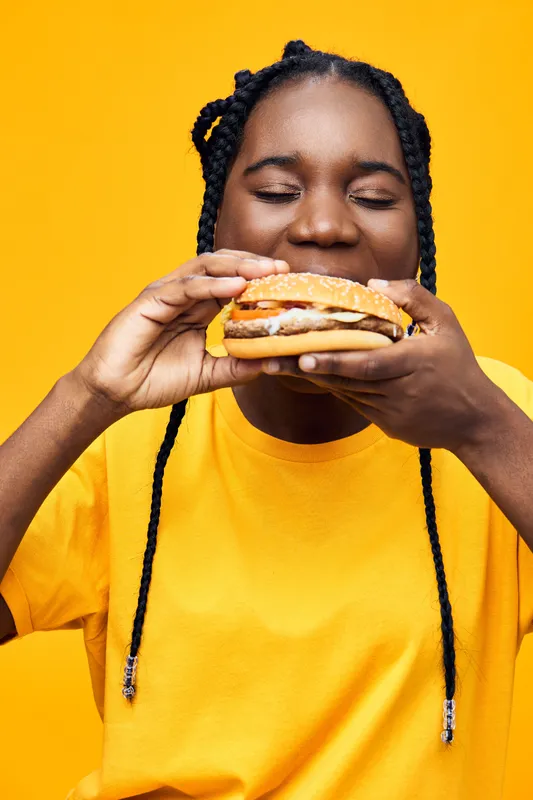 Happy young woman enjoying a delicious burger against a bright yellow background, showcasing her excitement and love for food, perfect for healthy eating concepts