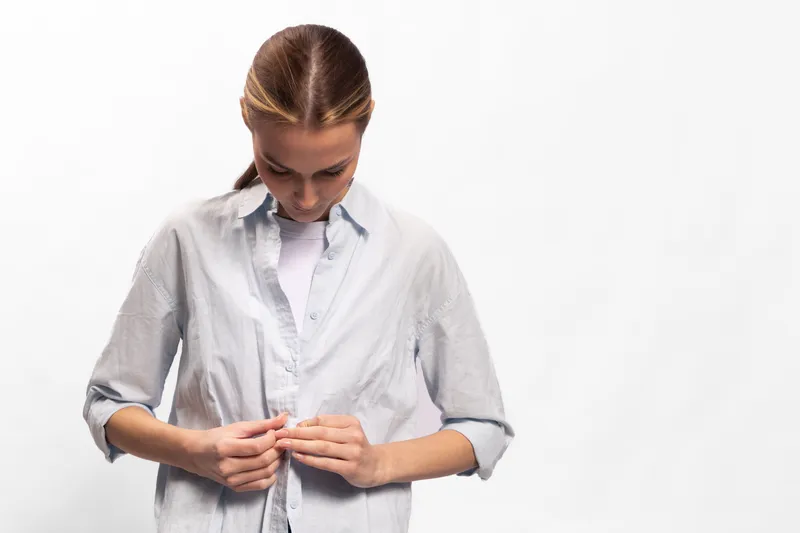 Young woman in casual shirt buttoning up, looking down. Minimalist studio on white background. Concept for clothing, fashion, daily routine, and casual style.