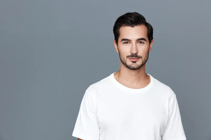 Confident young man with a stylish haircut, wearing a plain white t-shirt, posing against a solid gray background, exudes a sense of calm and approachability.