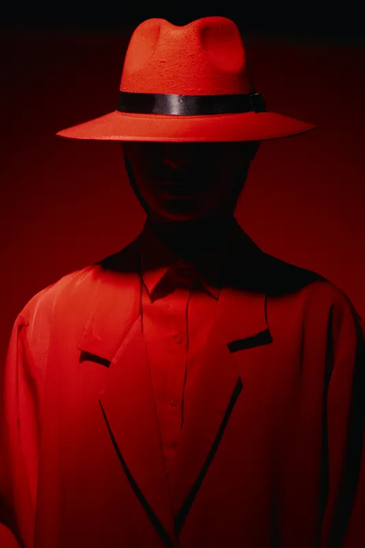 Mysterious person wearing a red fedora hat and red suit jacket stands against red background. Dramatic fashion lighting highlights the silhouette and creative mood.