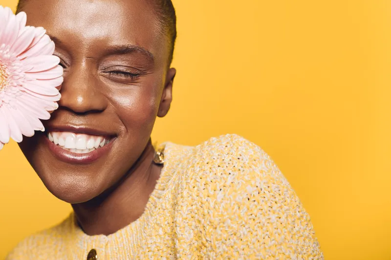 Close-up of happy dark skinned African woman with short hair smiling with closed eyes holding pink flower near face on yellow background. Concept of joy, beauty, and natural skincare.
