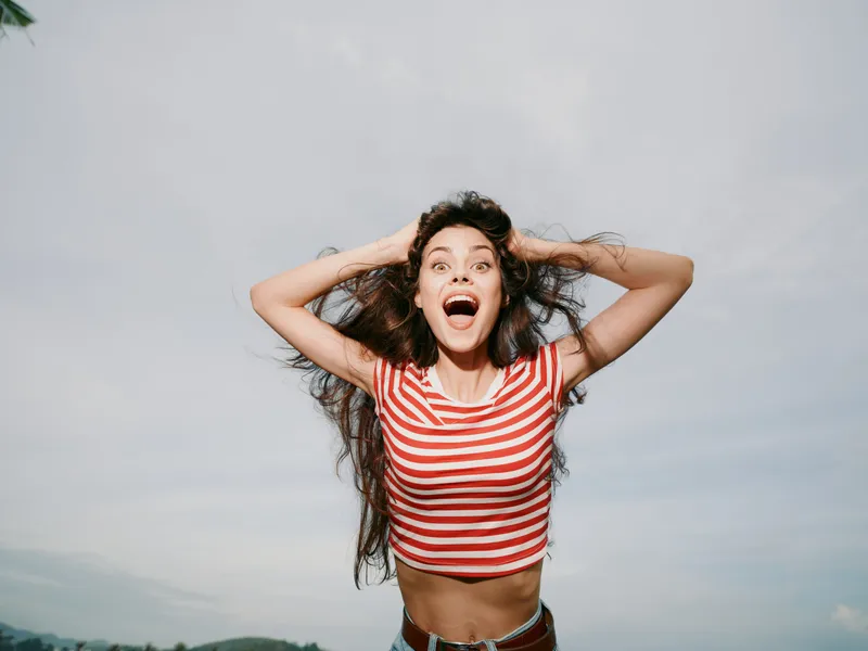 A joyful young woman wears a red and white striped top and stands against a bright sky, hands in her hair, radiating energy and carefree confidence.