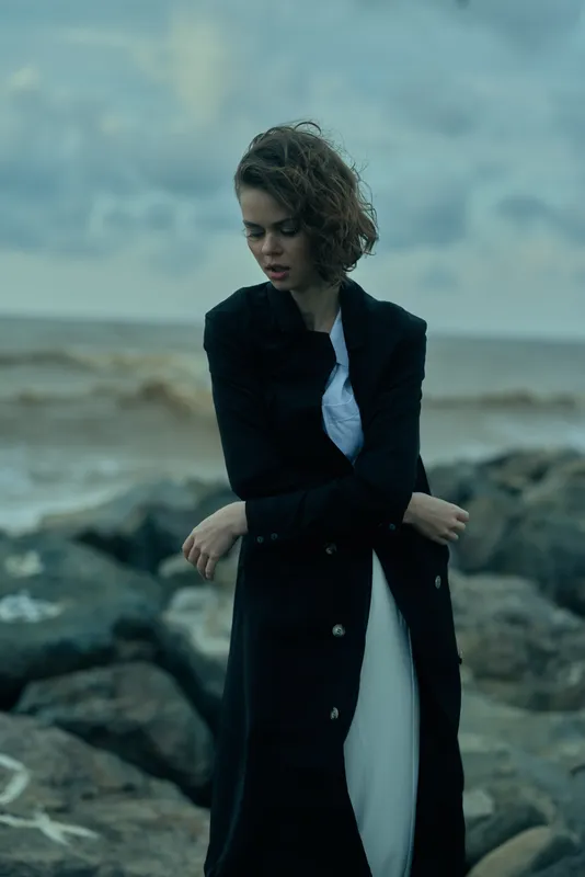 A woman in a dark coat stands on a rugged shoreline by the sea, looking down with a moody, contemplative pose amid windy coast