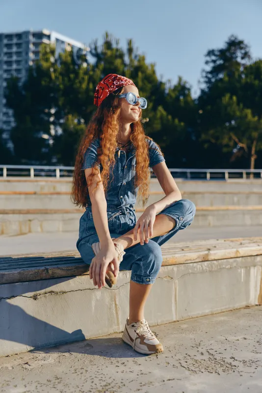lifestyle woman in modern boho-western denim street style sitting outdoors in warm sunlight with sunglasses and red bandana, film color ambiance