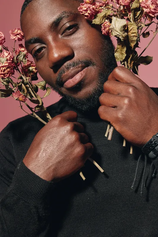 African man with dark skin holding dried flowers close to face in creative conceptual visual style. Portrait with emotional expression and artistic composition on pink background.