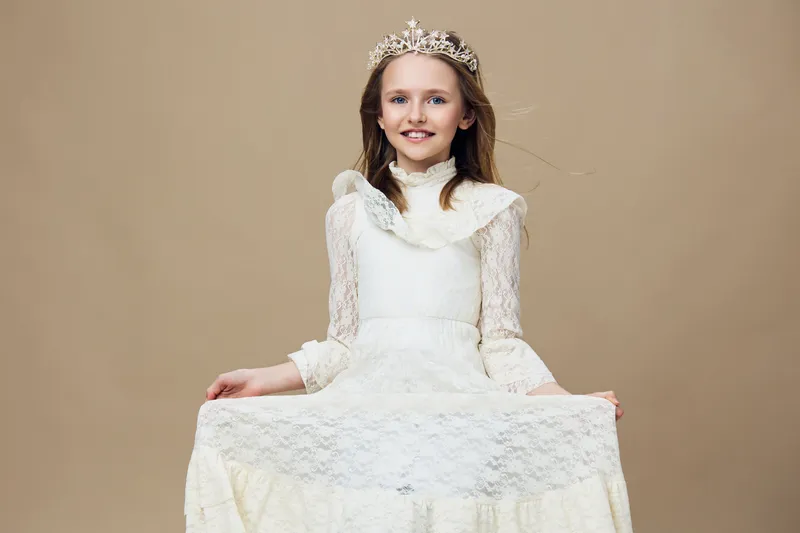 Young girl with a crown wearing a white lace dress against a neutral background, expressing joy and confidence in a timeless portrait style