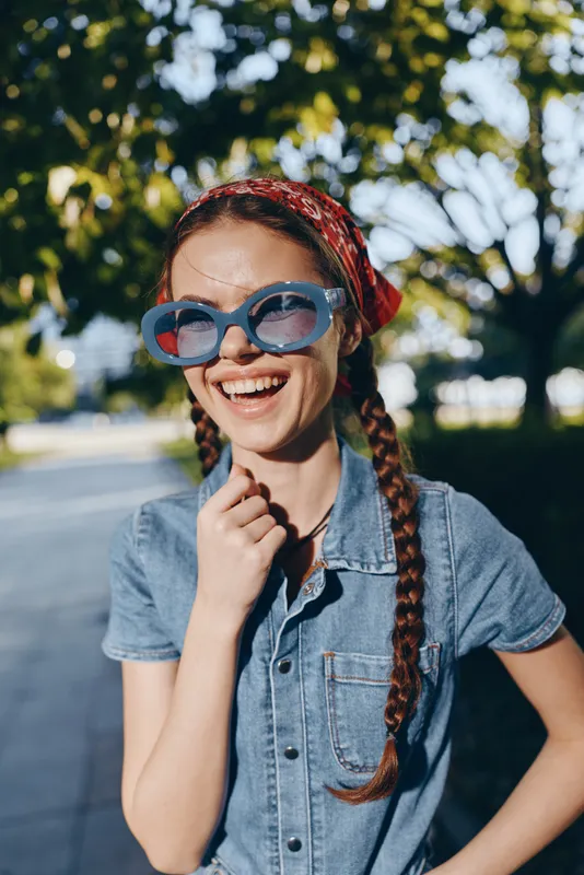 Lifestyle woman in modern boho-western denim street style smiles wearing oversized sunglasses and a red bandana, captured with warm film color tone outdoors.