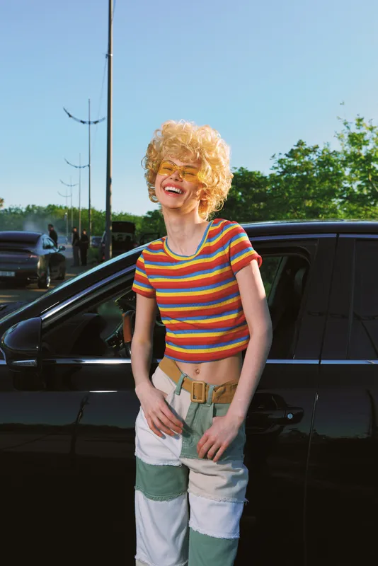Vibrant woman with curly blonde hair leans beside a black car in bright sun, wearing a rainbow striped tee and patchwork jeans. A playful retro lifestyle moment for photography.