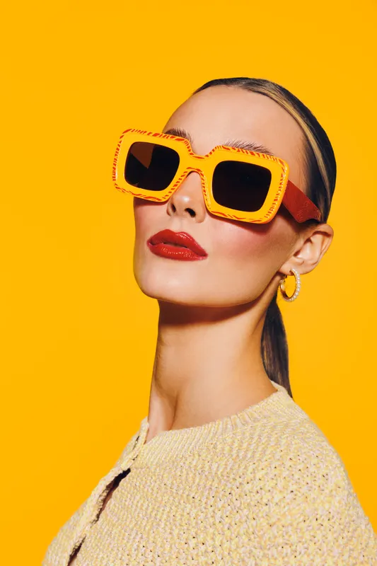 Stylish woman with slicked hair wears large orange sunglasses and red lipstick against bright yellow background. Fashion portrait with bold colors and confident expression.