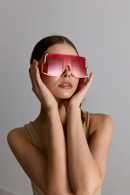 Young woman wearing oversized red sunglasses poses with hands framing her face, showcasing modern fashion accessory in minimalistic neutral setting with soft lighting.