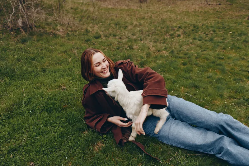 A young woman rests on grass, cuddling a goat kid in a brown coat, enjoying a peaceful outdoor moment. This lifestyle scene captures companionship, warmth, and gentle animal bonding in nature, with