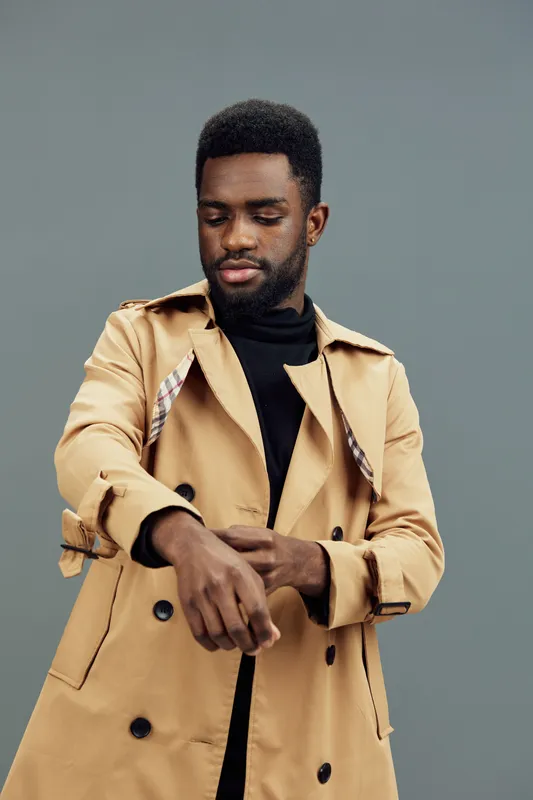 Stylish African man in a beige trench coat adjusting his sleeve, exuding confidence against a soft green background, showcasing modern fashion trends and seasonal elegance
