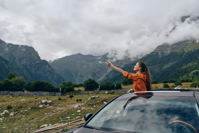 Adventure travel scene with a woman standing on a car roof, pointing toward distant mountains in a scenic landscape, capturing a bold carefree moment outdoors