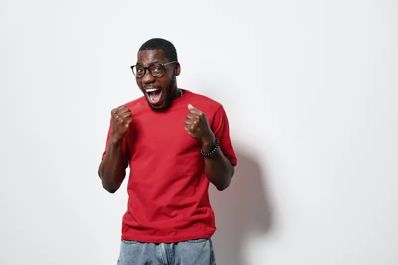 Excited African American man with dark skin wearing glasses and red t shirt celebrating success with clenched fists on white background. Happy young black male expressing joy.