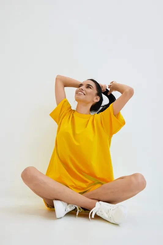 smiling woman in yellow dress sitting cross-legged on white floor, casual outfit, relaxed pose, bright studio background, youthful and joyful mood