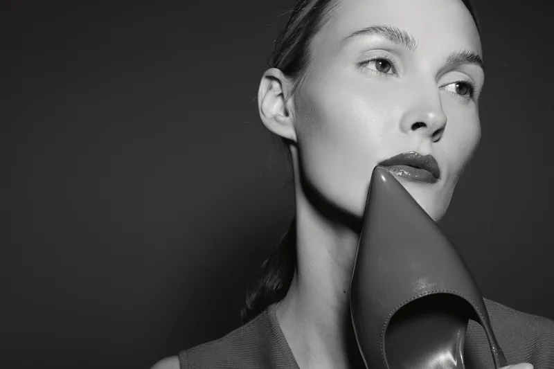 White woman with pale skin holds high heel shoe close to face in black and white portrait. Concept for fashion, beauty, and style with elegant mood.