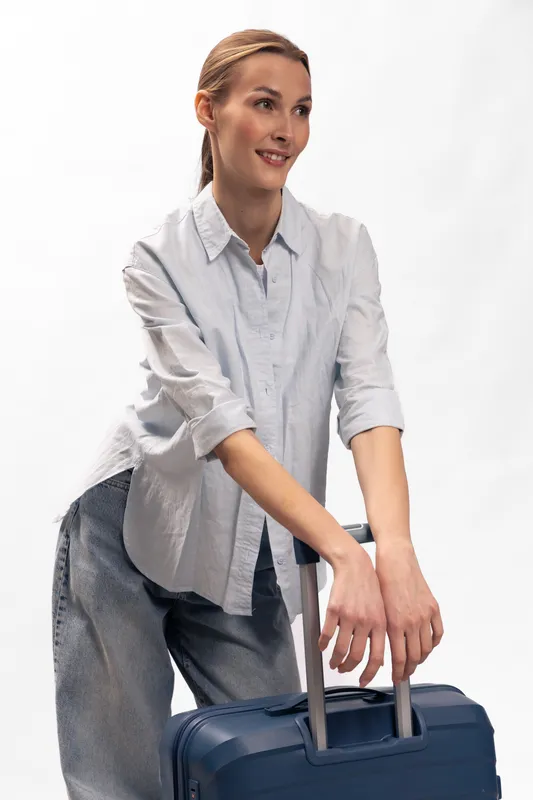 Young woman with light hair wearing casual shirt and jeans stands with blue suitcase on white background. Concept of travel, vacation, and preparation for trip.