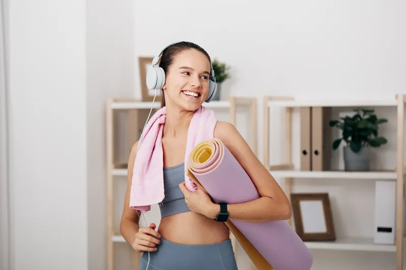 Fitness, sport, woman, workout in a bright indoor studio. She wears headphones, holds a rolled yoga mat and towel, and smiles, ready for a focused exercise session and home fitness training.