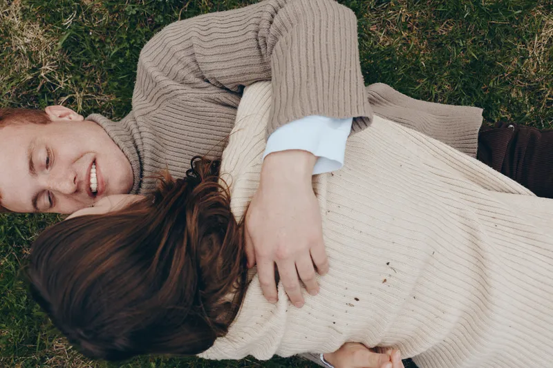 Two cheerful lovers lie on green grass embracing each other in cozy knit sweaters, enjoying a warm outdoor moment, romantic, relaxed lifestyle scene with sunlight and serenity in view