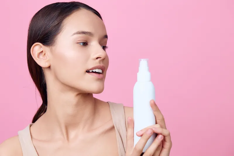 Young woman holding a white spray bottle, showcasing skincare, with a soft pink background, reflecting beauty, skincare routine, and self care concepts