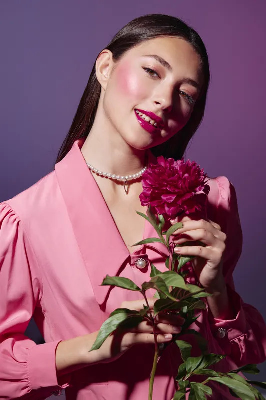 Young woman with vibrant makeup wears a pink blazer and pearl necklace, holding a deep pink flower. Artistic studio beauty fashion concept on a purple plain background.