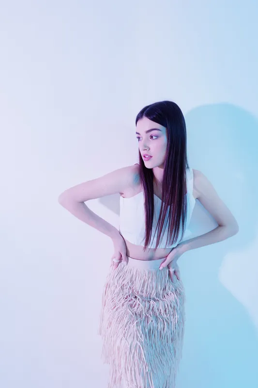 Young woman in a white crop top and textured skirt poses with hands on hips against a pastel background. Fashion concept focusing on beauty and style.