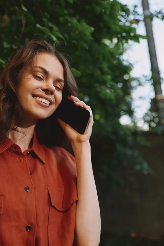 Cheerful woman outdoors on a phone, smiling with eyes closed, sunlit moment, relaxed posture, casual shirt, natural colors, lush greenery background, lifestyle vibe