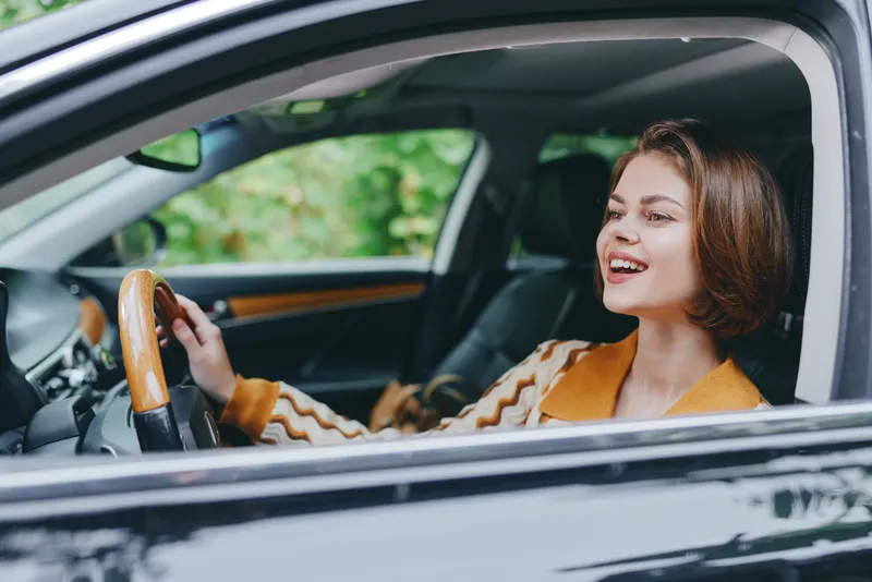 Joyful woman driving a car, eyes ahead, hands on the wheel, warm smile, stylish interior with wood accents and lush greenery outside.