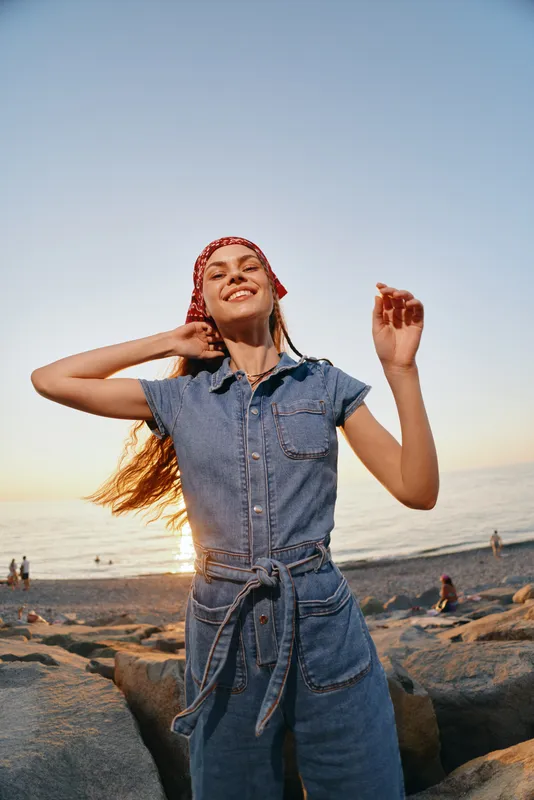 smiling woman in denim jumpsuit enjoys sunset on rocky beach with joyful expression and relaxed pose, golden hour lighting, clear sky background
