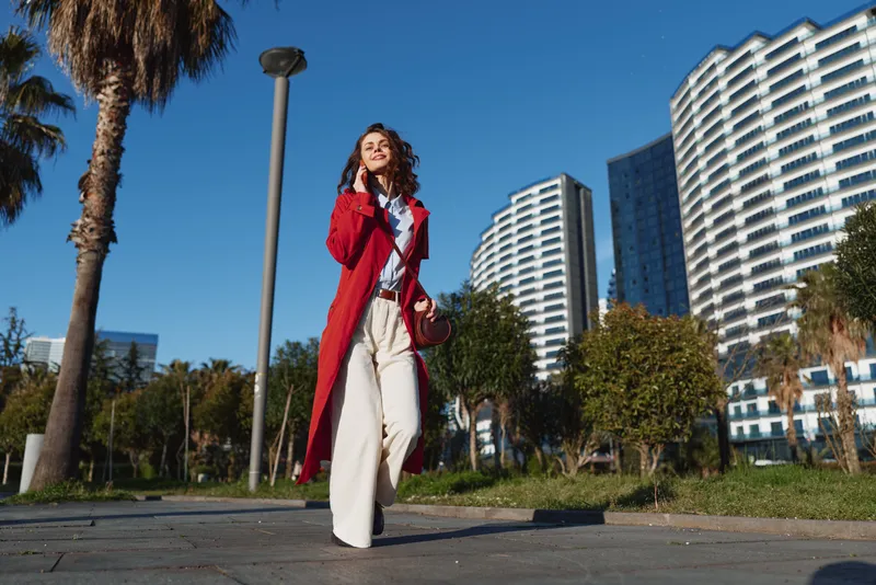Woman in a red coat walks outdoors in a cityscape, sunny daylight, smiling, fashionable pose, modern high rises behind, energetic urban style and confident vibe