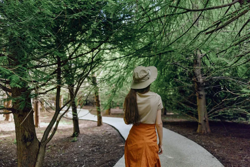 Woman walking on a curved path in a green forest, wearing a wide hat and orange skirt. Nature trail, trees and peaceful outdoor scene with back view and summer mood.