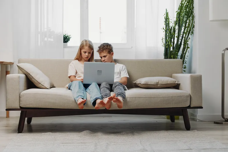 Two children sitting barefoot on a beige sofa using a laptop together in a bright living room with natural light and plants. Concept of technology, learning, and family bonding.