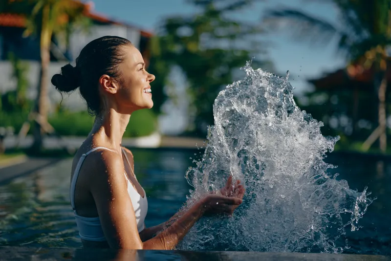 Cheerful woman in a pool outdoors, enjoying a sunny moment, water splash nearby, relaxed casual lifestyle vibe amid green garden scenery