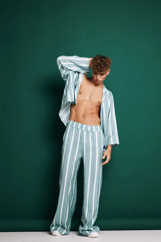 Young man with curly hair wearing striped pajamas standing against green background with shirt open showing toned abs and relaxed pose in casual indoor setting.