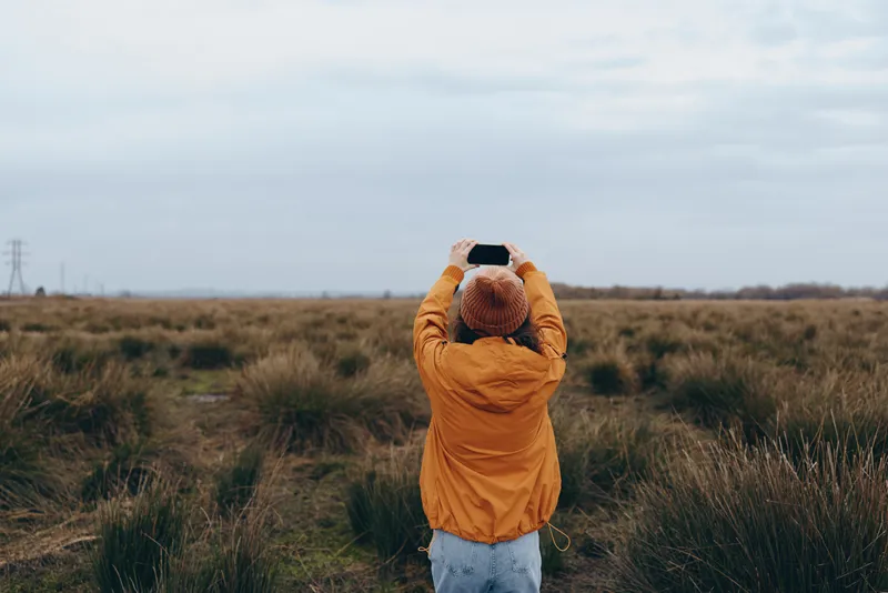 Woman outdoors lifestyle, standing in grassy field, orange jacket, knit hat, holding phone, looking at cloudy sky, calm traveler mood, natural light, wide landscape, soft breeze, autumn air