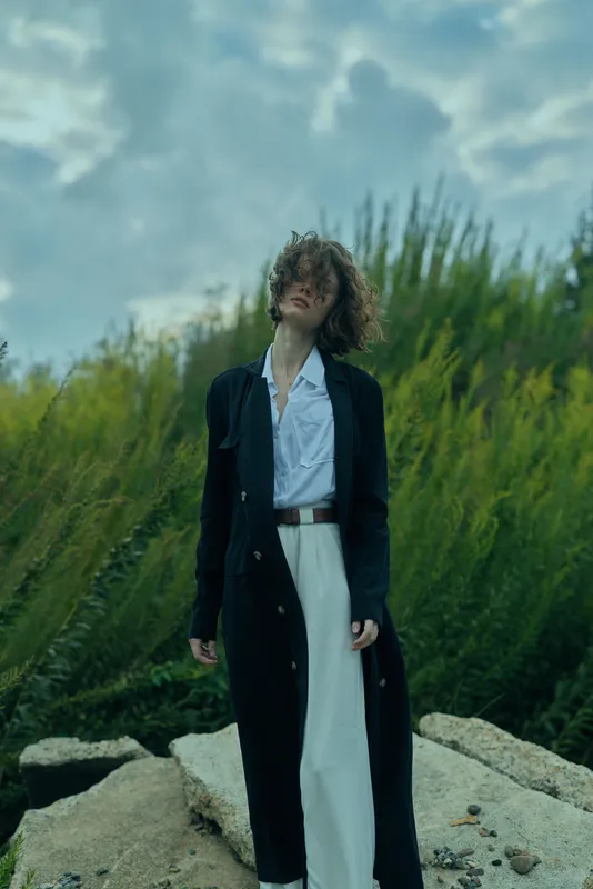 Woman in a long black coat over a white shirt and wide pants stands among rocks and reeds, wind in her hair, showcasing contemporary fashion and elegance