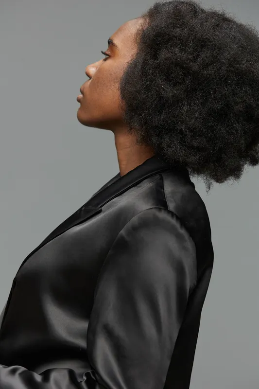 Elegant profile of a Black woman with natural hair, wearing a black satin blazer against a soft gray background, embodying confidence and sophistication