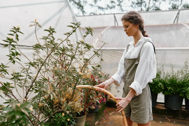 Young woman wearing overalls and a white shirt watering plants in a greenhouse with a serene expression, emphasizing the concept of sustainable gardening and nurturing nature