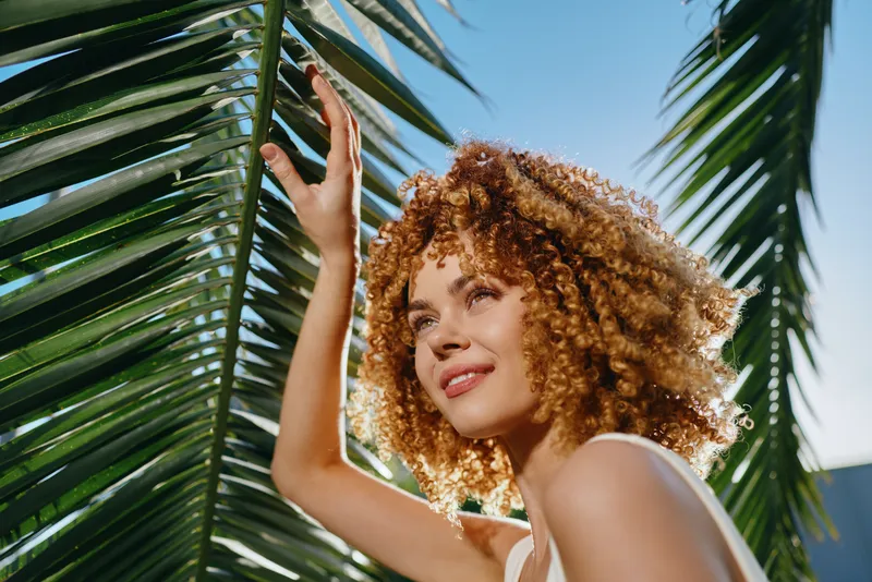smiling woman with curly hair enjoying sunlight under tropical palm leaves against clear blue sky in summer atmosphere
