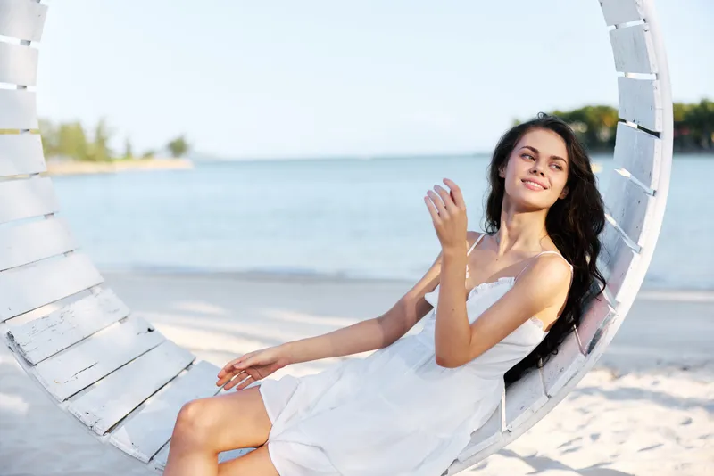 A woman sits on a circular white swing by the seaside, wearing a light dress, bathed in soft sunlight, with calm blue water and sandy shore creating a serene summer scene.