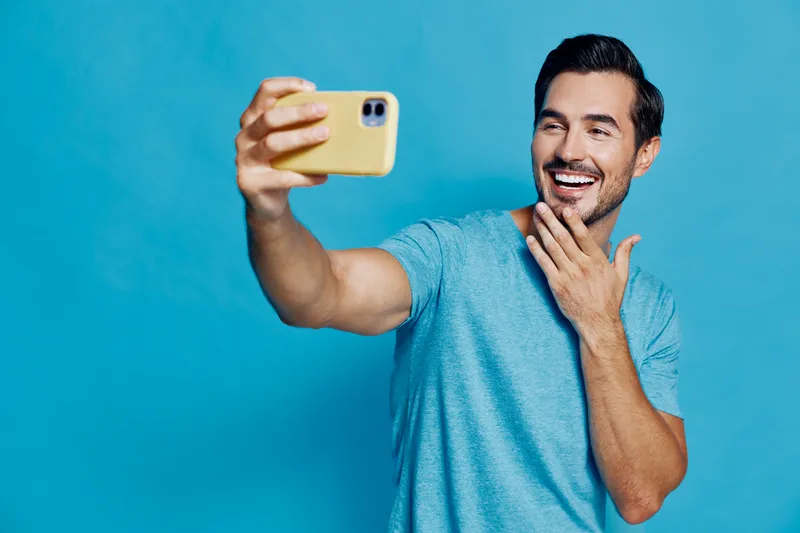 Smiling young man in casual blue t-shirt holding smartphone taking selfie on bright blue background, showcasing a joyful moment of self-expression. People lifestyle concept.